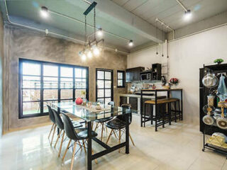 Modern kitchen and dining area with a glass-top table, black chairs, bar-style seating, hanging lights, large windows, and wall-mounted shelves. Bright, spacious interior with a mix of industrial and contemporary design elements.