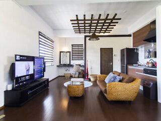 A modern living room with a flat-screen TV, wicker furniture, hardwood floor, small kitchen area, and minimalist decor.