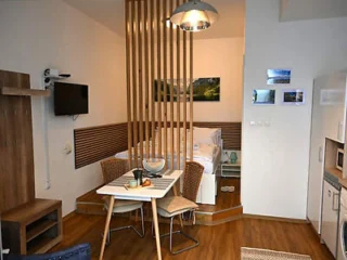 A small studio apartment featuring a bed, a partition with vertical wooden slats, a dining table with two chairs, a wall-mounted TV, and a kitchenette.
