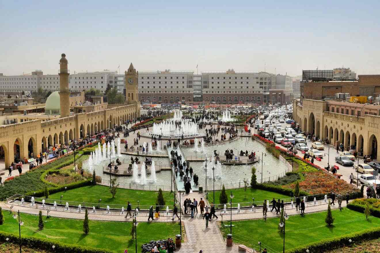 A bustling square with fountains and green spaces surrounded by crowded streets and historic buildings. Many people are walking and socializing.