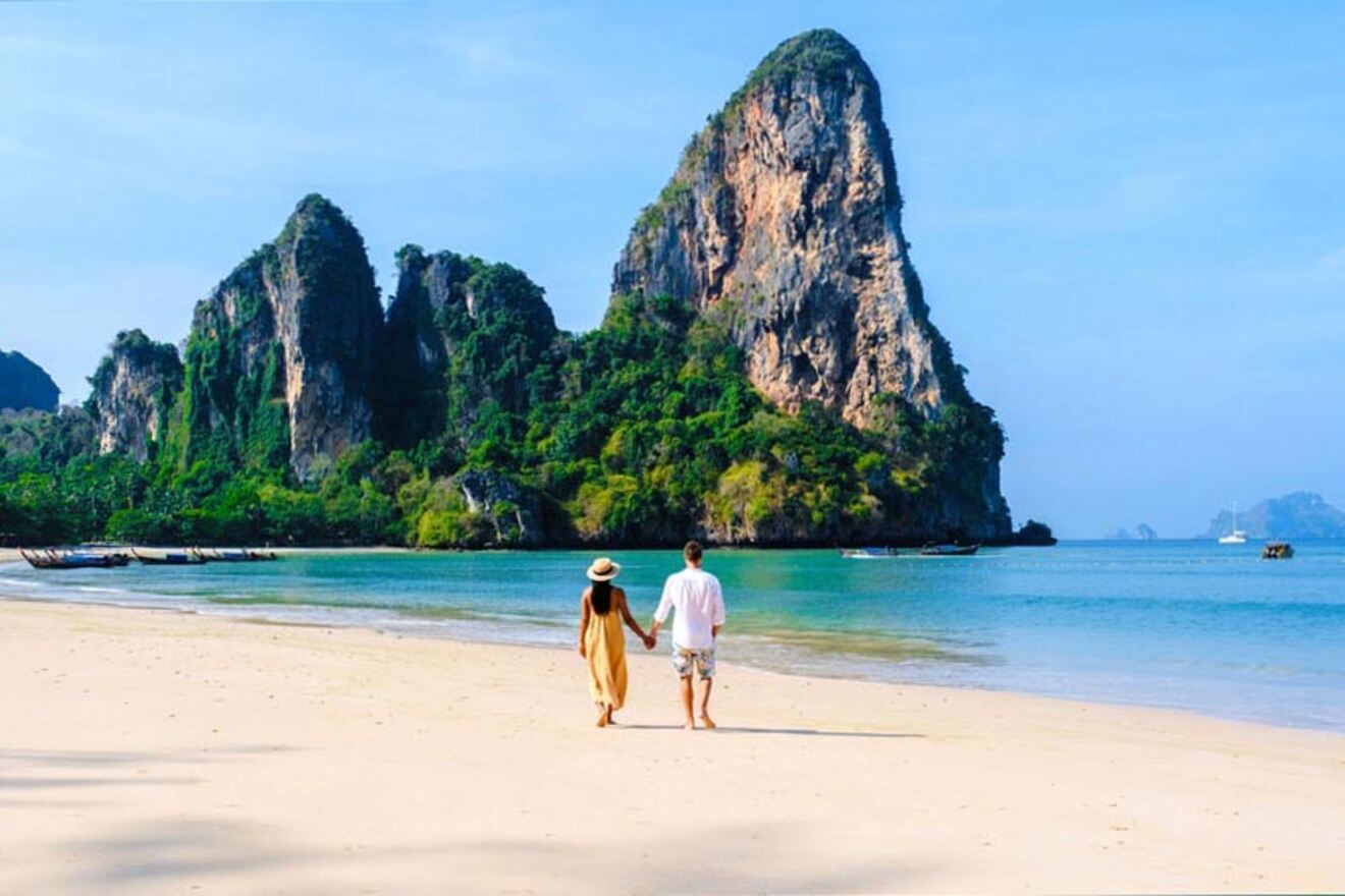 A couple holding hands walking on a sandy beach towards limestone cliffs under a clear blue sky.