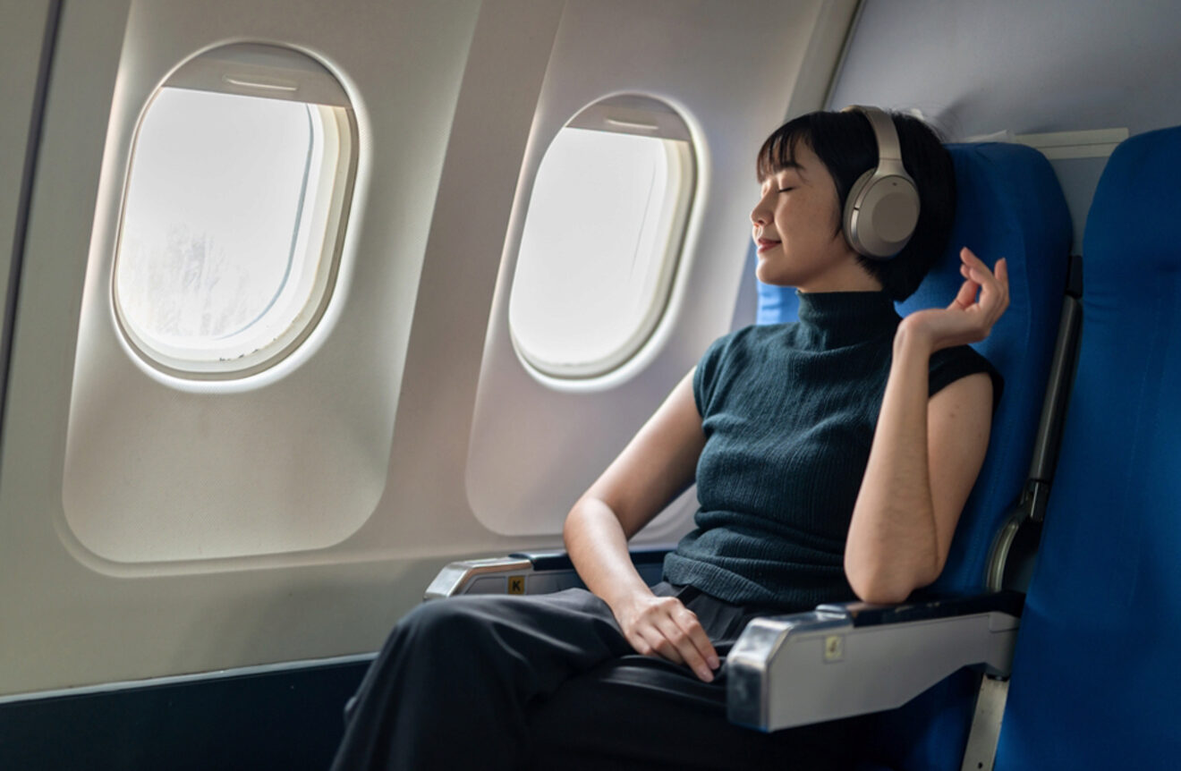 A woman with short hair, wearing large headphones, relaxes with eyes closed in an airplane seat.