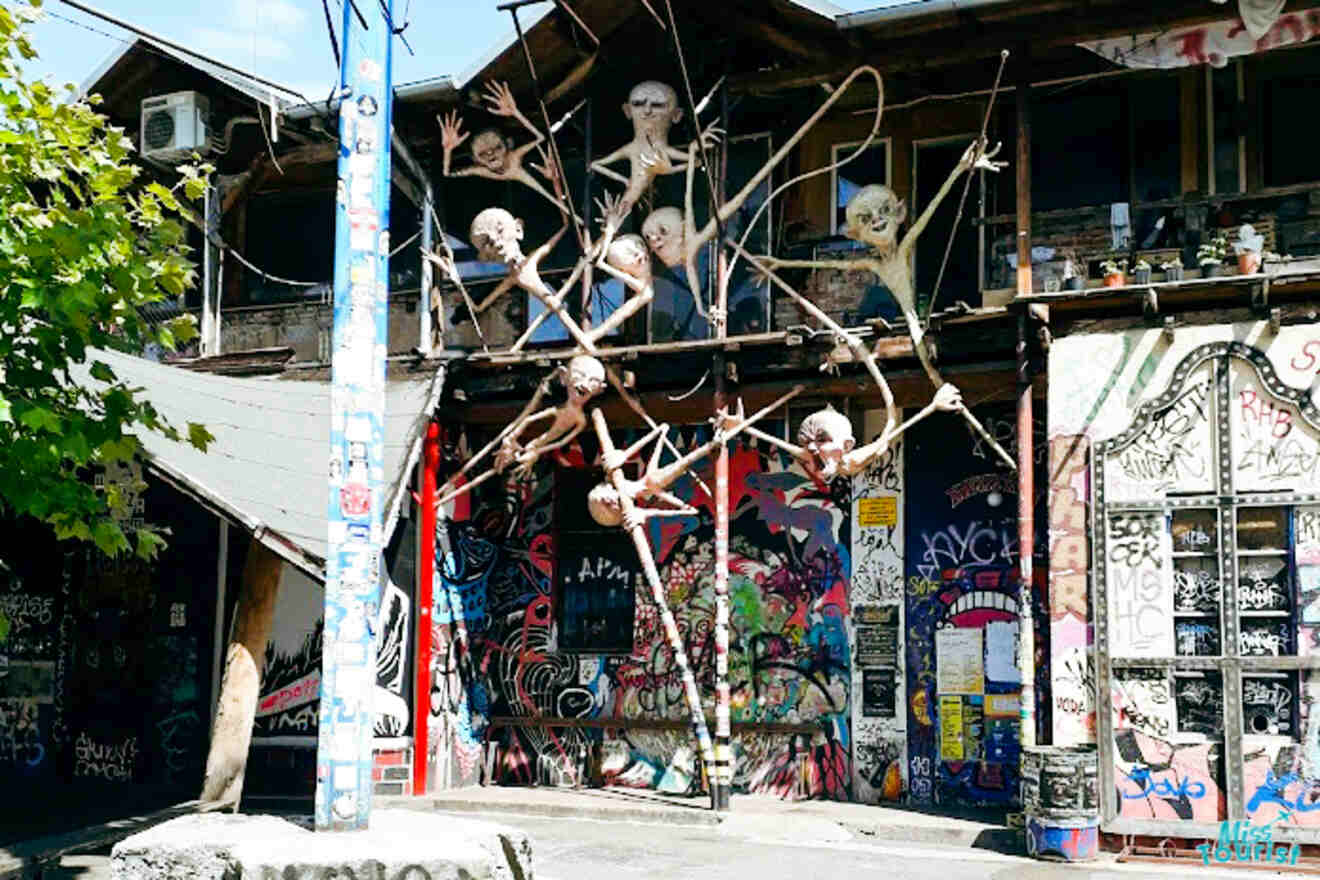 A graffiti-covered building featuring multiple eerie sculptures clinging to its exterior, with tree branches and a mix of human and creature-like figures.