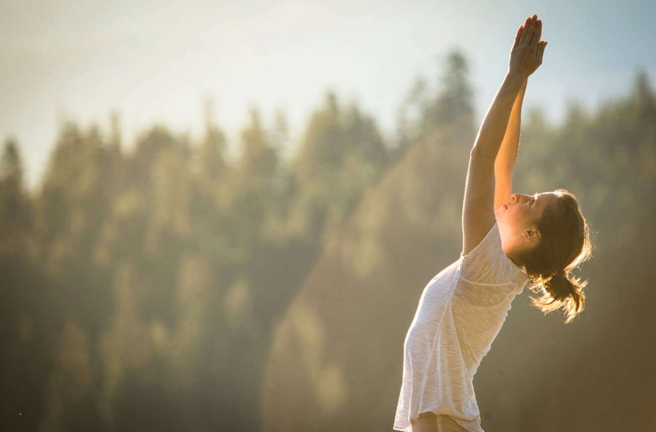A woman practicing a yoga stretch outdoors, reaching her arms upwards with a forest background in the early morning light
