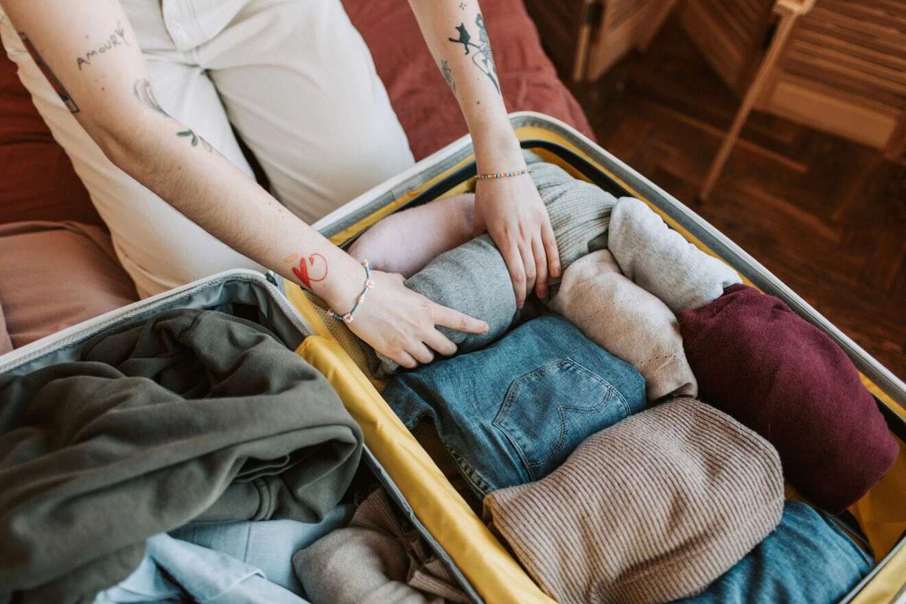 A person is packing or unpacking a suitcase filled with neatly folded clothing, including sweaters, jeans, and shirts. The person's arms have visible tattoos.