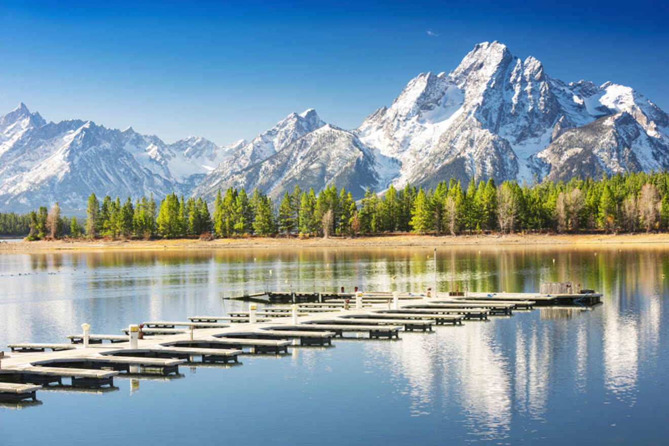 A serene lake with multiple wooden docks extends toward a backdrop of snow-capped mountains and a pine forest under a clear blue sky.