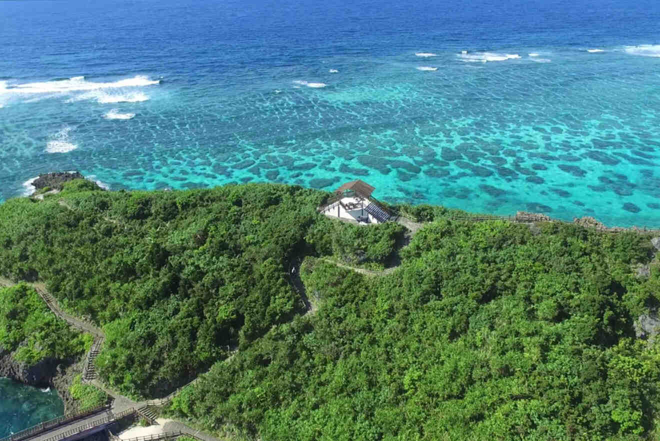 Aerial view of a small building surrounded by lush green vegetation on a cliff overlooking turquoise ocean waters with visible coral reefs.