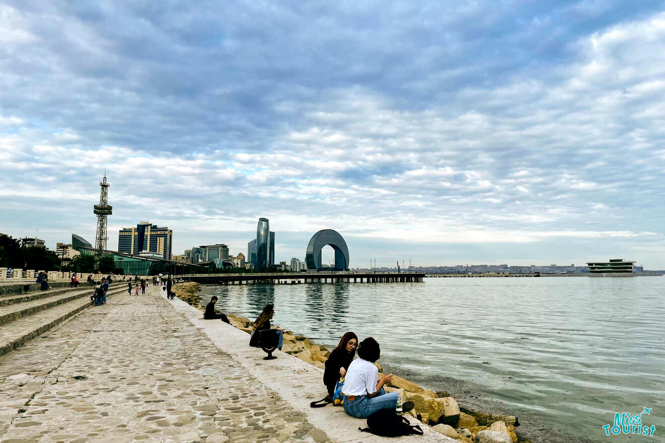 A few people sit and walk along a waterfront promenade with an urban skyline and a distinctive arch-shaped building in the background under a cloudy sky.