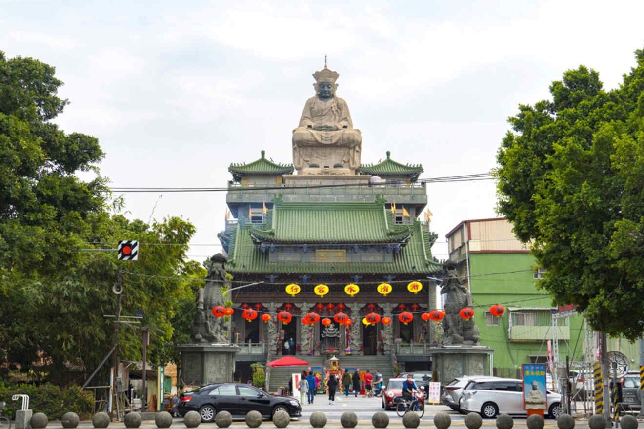 Large statue of a seated figure above a traditional building adorned with green roofs and red lanterns. Several people and cars are visible in the foreground.