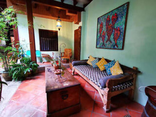 A covered patio with a wooden sofa, patterned cushions, a rectangular coffee table, and a framed artwork on the wall. There is greenery and a blue chair in the background.