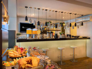 A modern café with a counter featuring hanging lights, bar stools, and an assortment of pastries and fresh fruits on the foreground table.