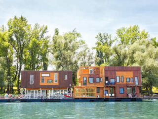 Two modern houseboats with wooden exteriors are moored on a calm river, surrounded by lush green trees under a clear sky.