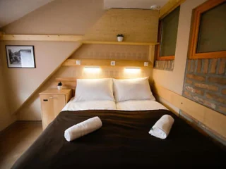 A cozy bedroom with a double bed, two rolled-up towels, a wooden nightstand, framed photo, recessed lighting, and a small plant on a shelf above the bed.
