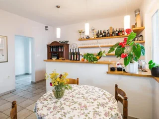 A cozy dining area with a round table covered by a floral tablecloth, surrounded by wooden chairs. Shelves with wine bottles and decorative plants line the wall behind a small bar counter.