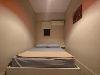 A small, tidy bedroom features a bed with blue sheets, a folded tan blanket, and two pillows. A closed window with a blind and an air conditioner are on the wall above the bed. There is a painting on the right wall.
