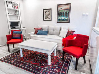 A small, modern living room with a white sofa, two red chairs, a white coffee table, a computer on a shelf, and two paintings on the wall. The floor is covered with a patterned red rug.