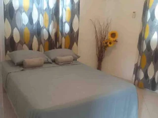 A neatly made bed with gray bedding is centered in a room, flanked by patterned curtains featuring gray and yellow leaf designs. A vase with sunflowers stands in the corner beside the window.