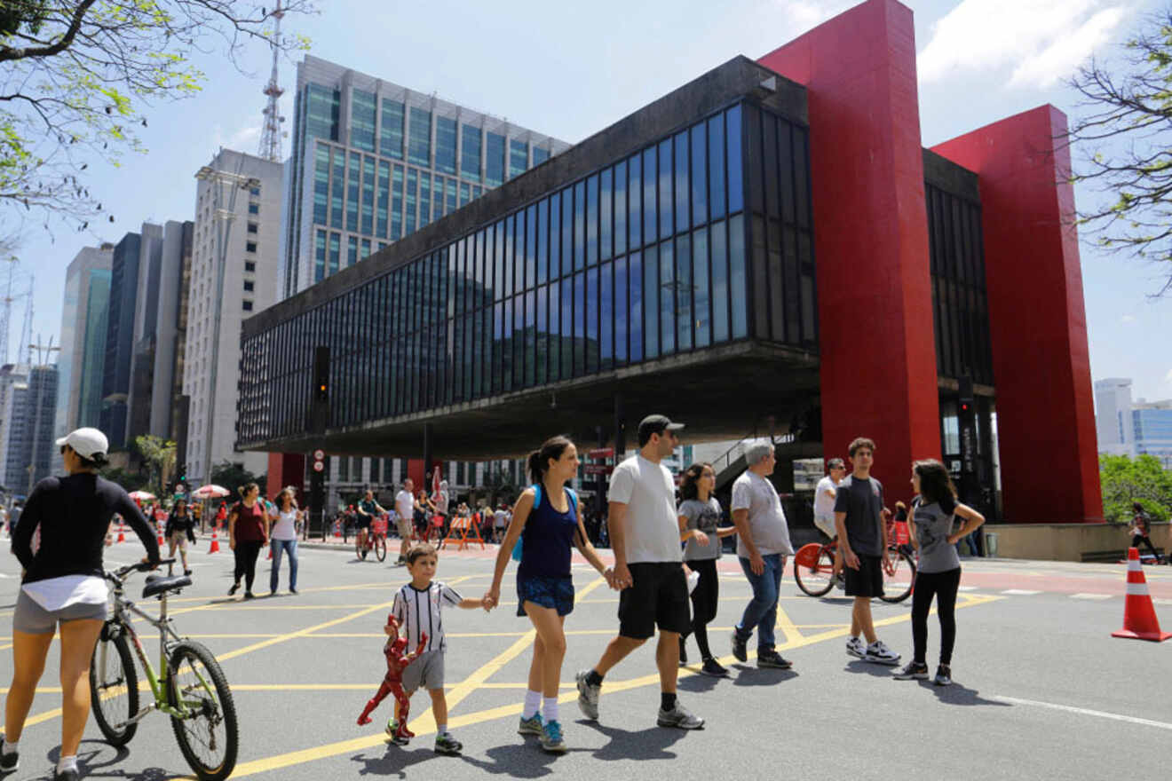 People walking and biking near a modern glass and red building in an urban area on a sunny day.
