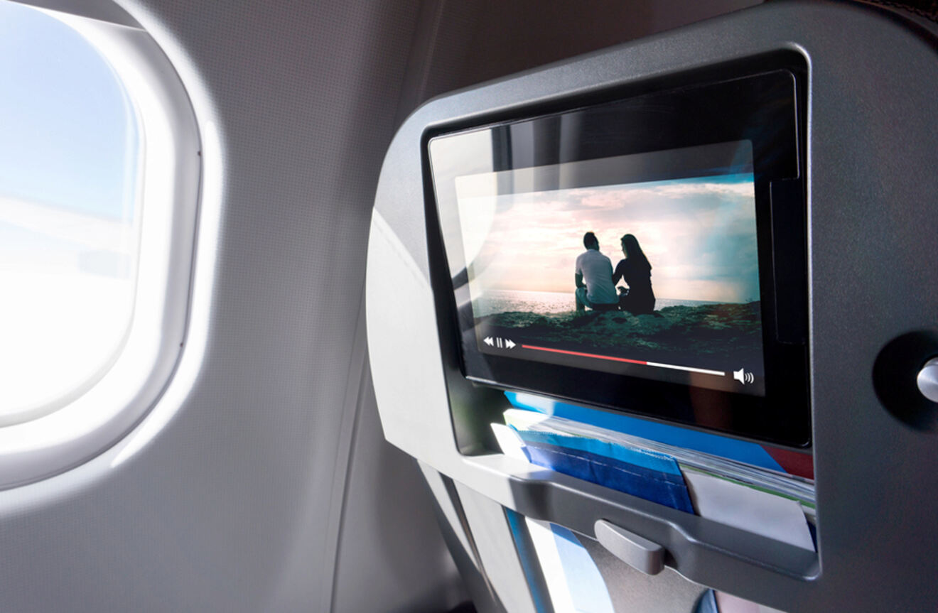 An in-flight entertainment screen displaying a movie scene of two people sitting on a beach, with a window beside it.