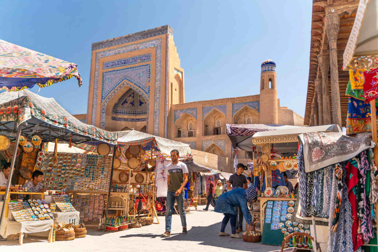 People browse at an outdoor market with colorful stalls selling various items. In the background is a large historical building with intricate blue tile work and an arched entrance.