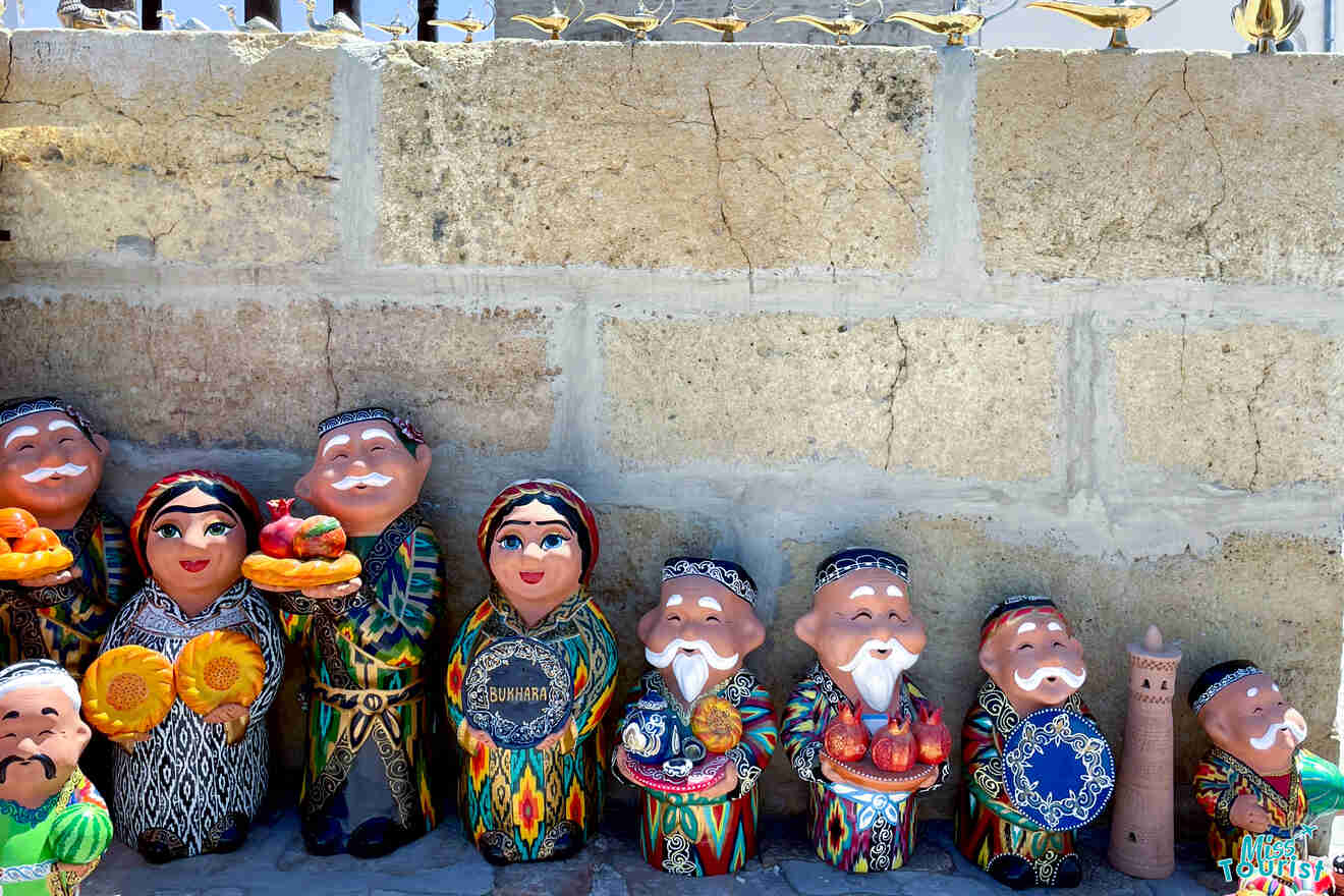 A row of colorful ceramic figurines, including men, women, and a child, dressed in traditional attire, holding various items like fruit and a cat. They are displayed on a stone ledge against a wall.