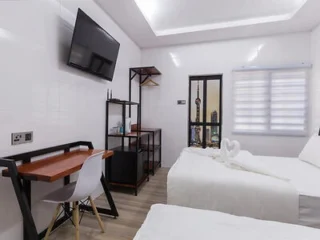 A modern, minimalistic hotel room with two white beds, a wall-mounted TV, a desk with a chair, and an open shelving unit beside the window with blinds.