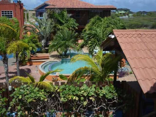 A small, tropical-style pool area surrounded by palm trees and vibrant greenery is overlooked by red-roofed buildings. In the background, a blue sky and distant landscape are visible.