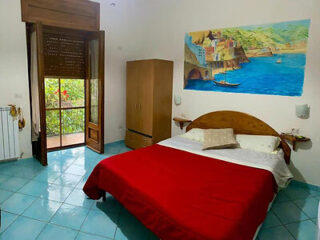 A bedroom with a large bed covered with a red blanket, a wardrobe, and a mural depicting a coastal scene. There is a door leading to a terrace with greenery outside. The floor is tiled in blue.