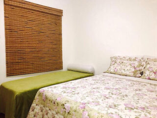 A bedroom with a floral-patterned double bed and a smaller green bed on the left. The window has bamboo blinds, and the walls are plain white.