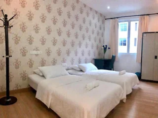 A room with two single beds, white linens, a standing coat rack, a desk with a chair, a small vase with flowers, and a large window with curtains. The walls are decorated with floral wallpaper.