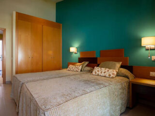 A bedroom with two single beds side by side, matching bedspreads, and headboards in front of a turquoise wall. There are two wall-mounted lamps above the beds and a closed wooden wardrobe on the left.