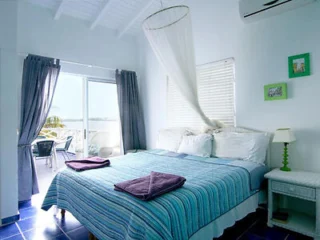A bright bedroom with a large bed, purple towels, striped bedspread, mosquito net, air conditioner, and sliding doors leading to an outdoor patio with seating.