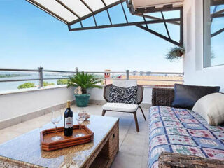 A rooftop terrace with a river view includes a wicker sofa and chair, a table set with wine, glasses, and a plant. The terrace is covered by a metal and glass canopy.