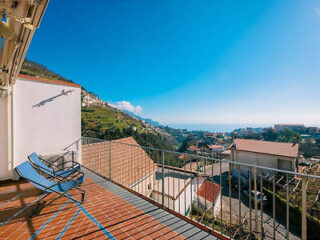 A balcony with two lounge chairs overlooks a hillside town and the sea under a clear blue sky.