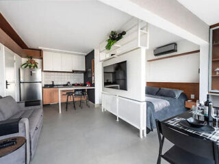 Modern studio apartment featuring a compact kitchen, a small dining area with two chairs, a bed, a couch, a wall-mounted TV, and minimalist decor with visible air conditioning unit and plants.