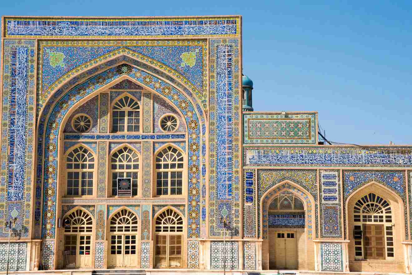 A brightly colored tiled facade of a mosque featuring intricate geometric patterns and arched windows, set against a clear blue sky.
