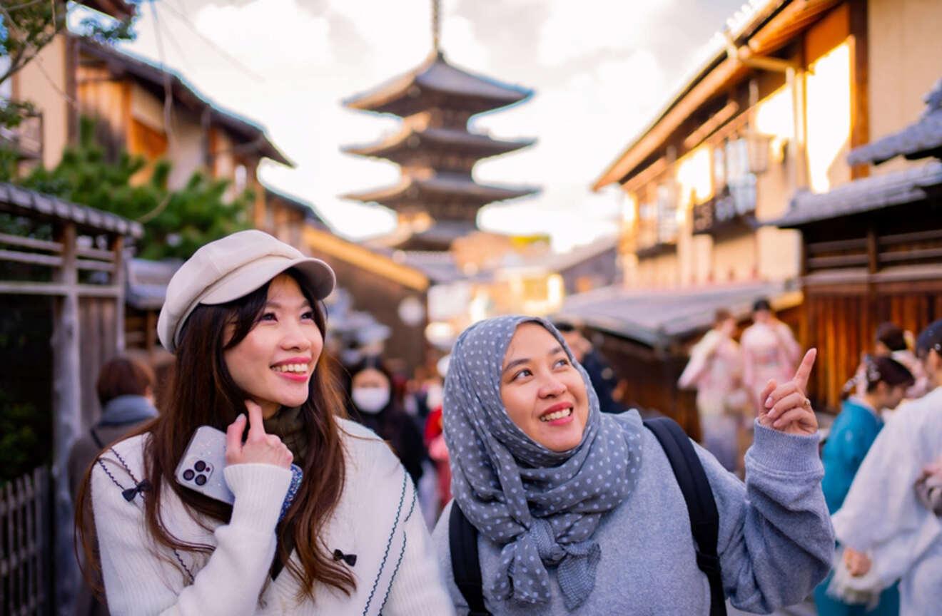 Two women happily explore a traditional street with a pagoda in the background, one pointing at something.