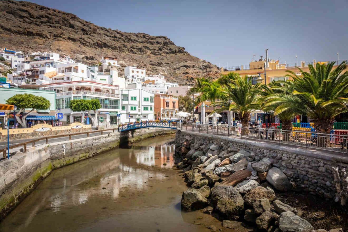 A scenic view of a coastal town featuring buildings on hills, a stone canal with clear water, a small blue bridge, and palm trees along the walkway on a sunny day.