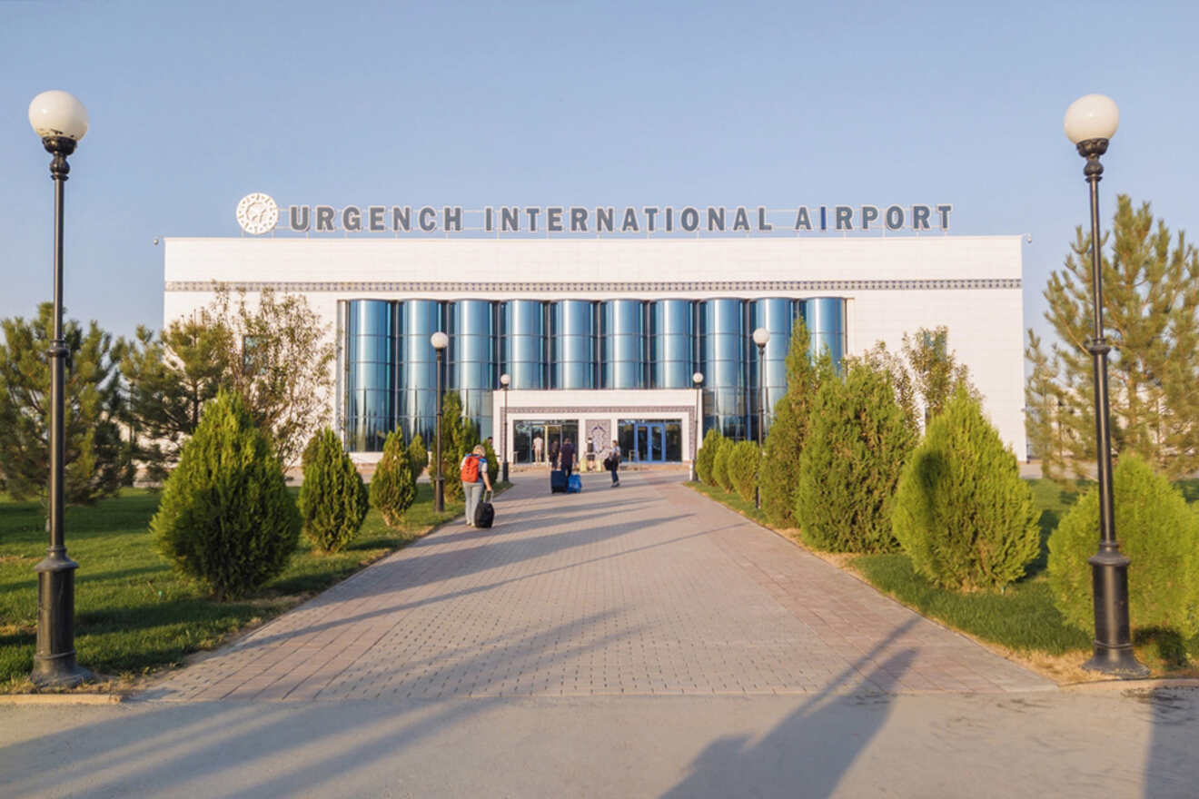 Front exterior of Urgench International Airport with two travelers walking along a pathway flanked by trees and lamp posts.
