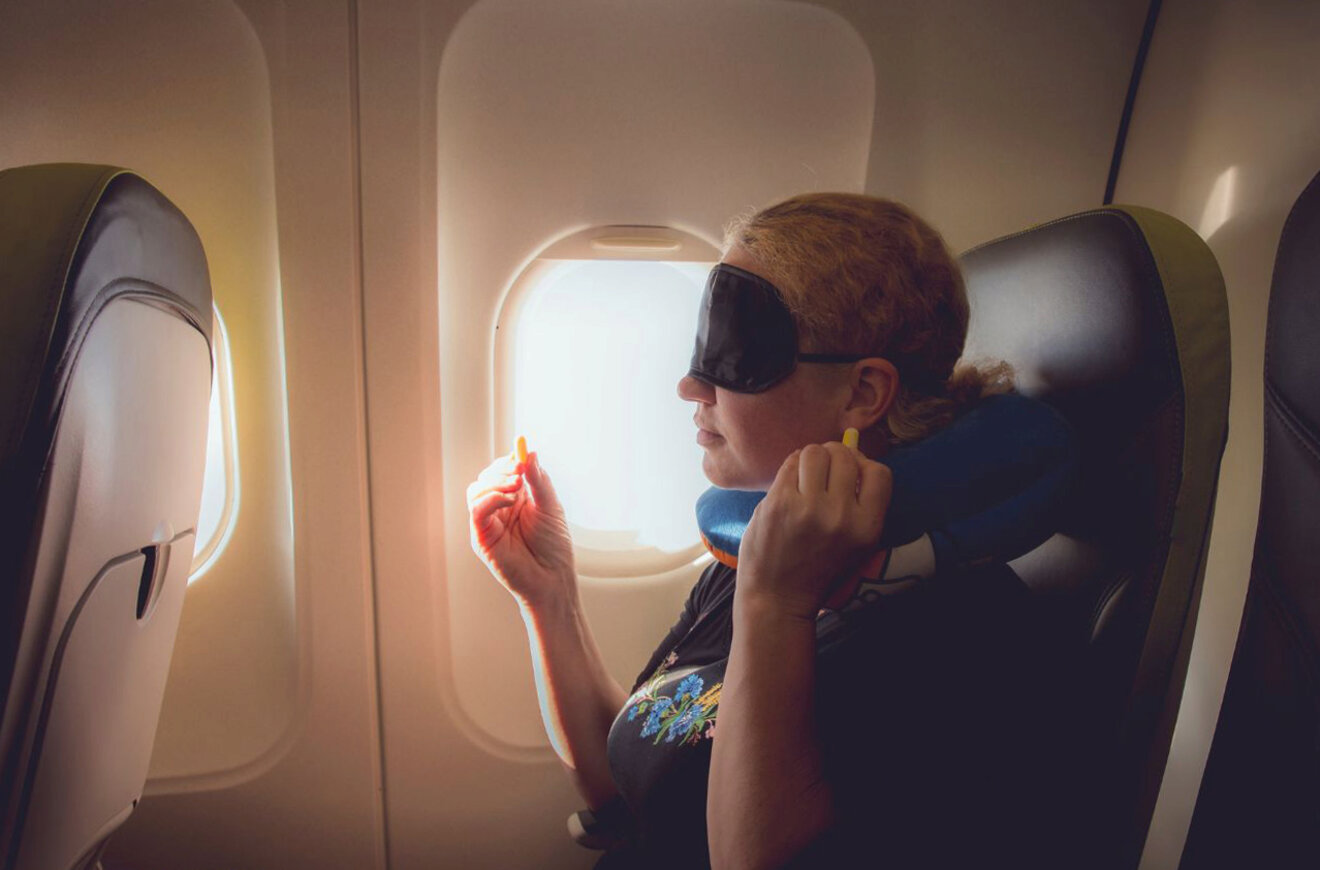 A woman with a sleep mask and neck pillow prepares to insert earplugs while seated on an airplane.