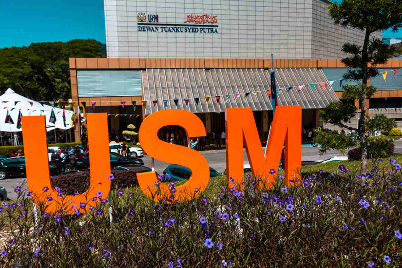Large orange "USM" letters in front of Dewan Tuanku Syed Putra building, surrounded by purple flowers and colorful flag decorations.