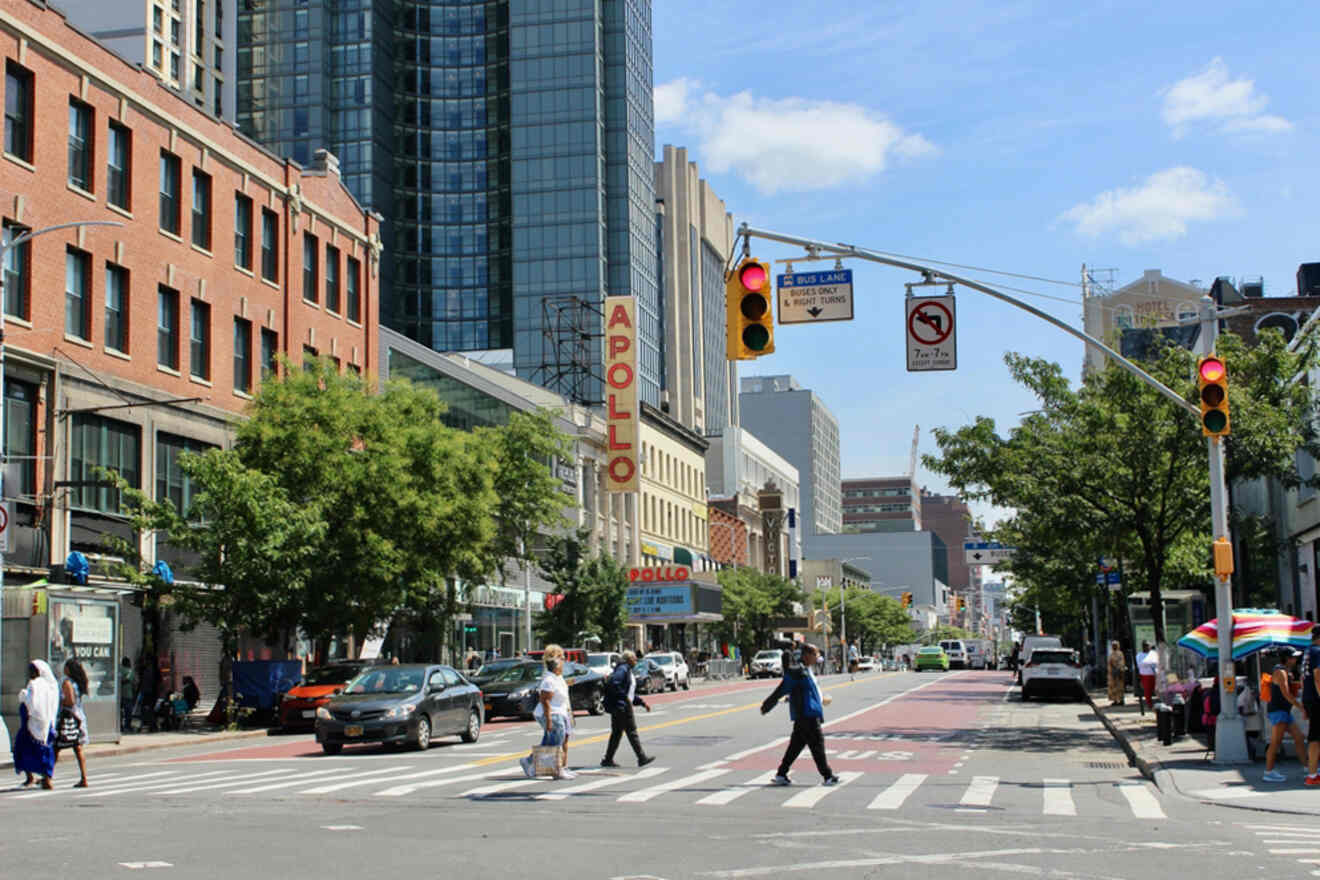 Street scene with pedestrians crossing a crosswalk near the Apollo Theater in an urban area. Buildings, traffic lights, and cars are visible under a clear sky.