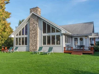 A gray A-frame house with large windows and a stone chimney, set on a well-maintained grassy lawn with outdoor lounge chairs on the deck and lawn area.