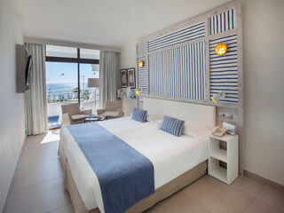 Modern hotel room with a large bed, blue and white decor, wall-mounted TV, and a seating area by a window overlooking a balcony and the ocean.