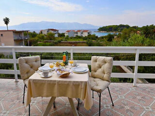 A small table set for two with breakfast items is on a balcony overlooking a scenic view of houses, greenery, and a distant body of water under a partly cloudy sky.