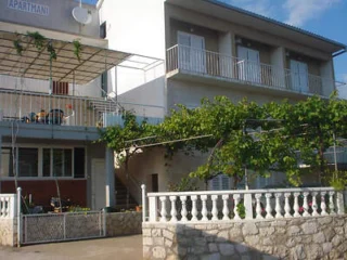 A two-story building with balconies and a vine-covered terrace. The ground floor has a red and white facade and a stone railing. There is a sign reading "APARTMANI" on the upper left corner.