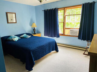 A bedroom with blue walls, blue curtains, a large window, a double bed with blue bedding and pillows, a bedside table with a lamp, and an air conditioning unit in the window.