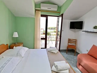 A bedroom with a double bed, bedside table with a lamp, TV mounted on the wall, an armchair, and a door leading to a balcony. Walls are green, and the room has folded towels on the bed.