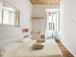 A minimalist bedroom with a double bed, two pillows, a pink bedside lamp, an open wardrobe, a window, and a small table with two chairs. The ceiling has a rustic brick design.