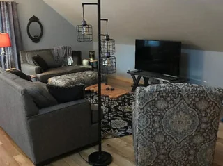 A living room with grey couches, patterned chairs, a TV on a stand, a wooden coffee table, a lamp with cage-like shades, and a mirror on the wall.